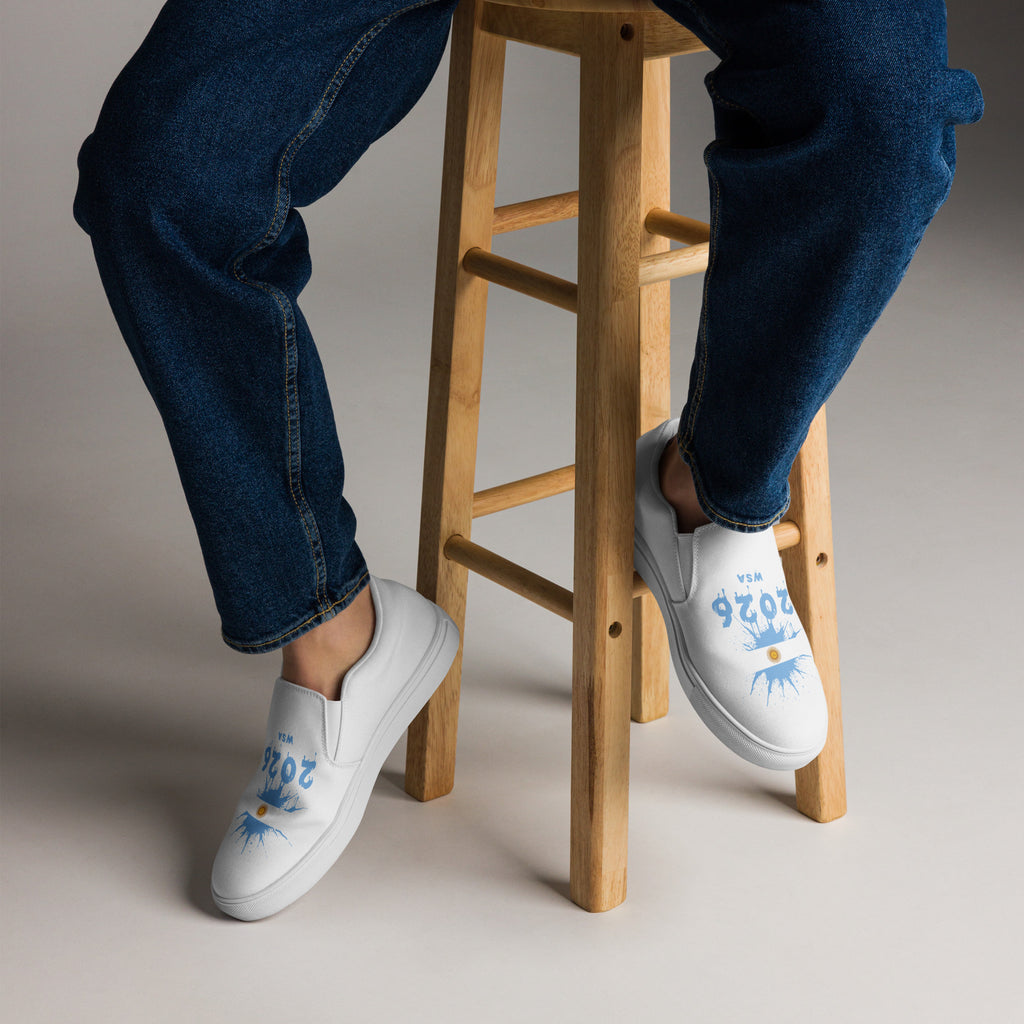 White sneakers with blue designs worn by a person sitting on a wooden stool.
