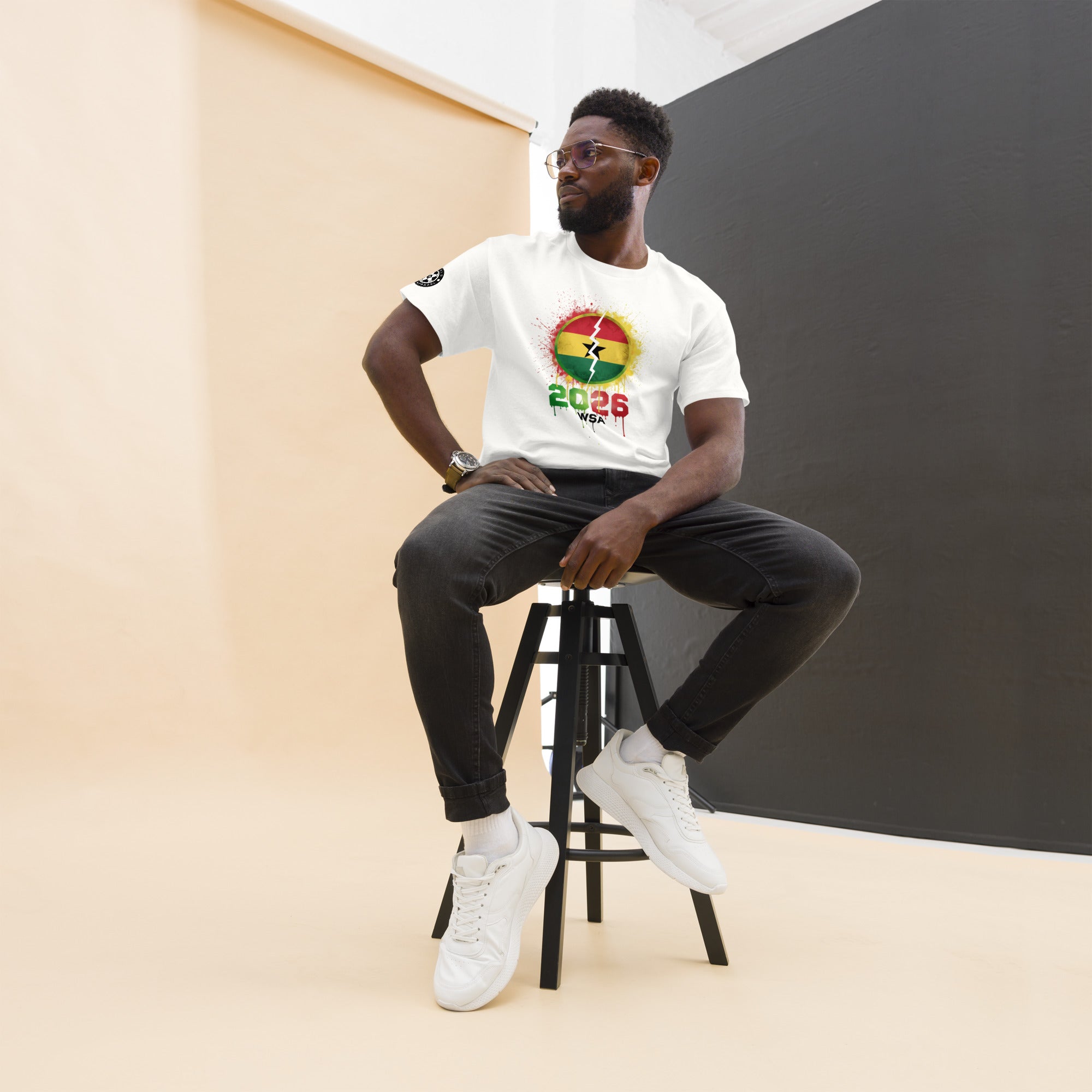 Man sitting on a stool wearing a white t-shirt with a colorful design, black pants, and white sneakers.