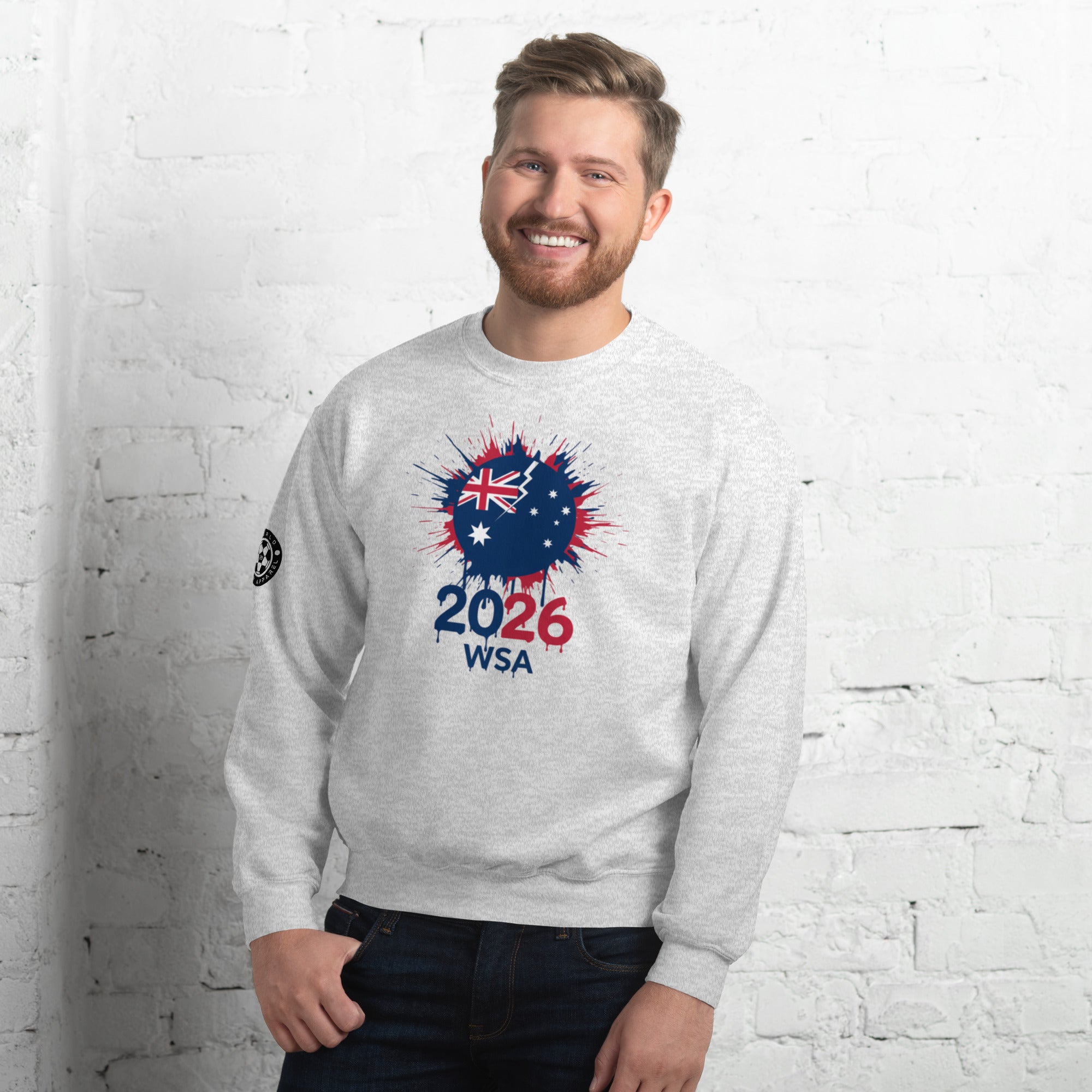 Man wearing a gray sweatshirt with a design featuring the number 2026 and text, standing against a white brick wall.