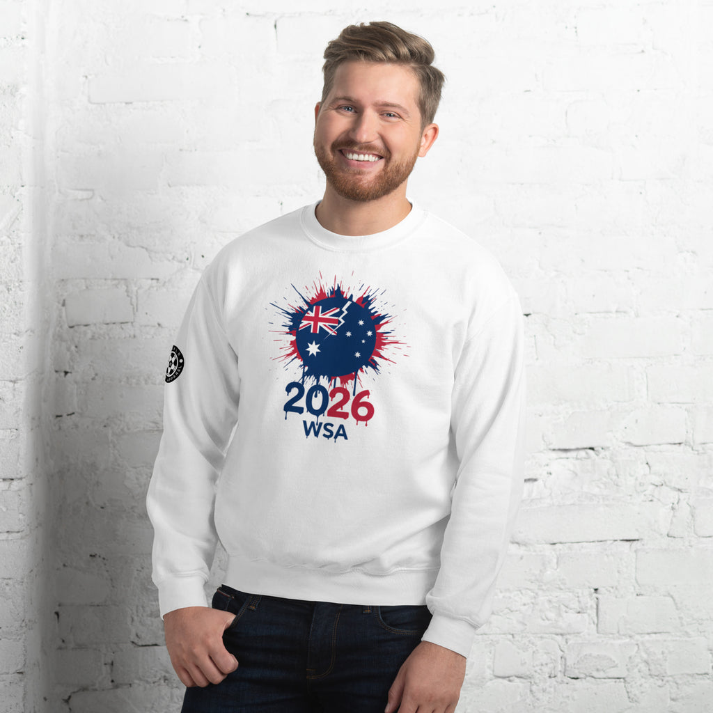 Man wearing a white sweatshirt with a design featuring the number 2026 and text, standing against a white brick wall.