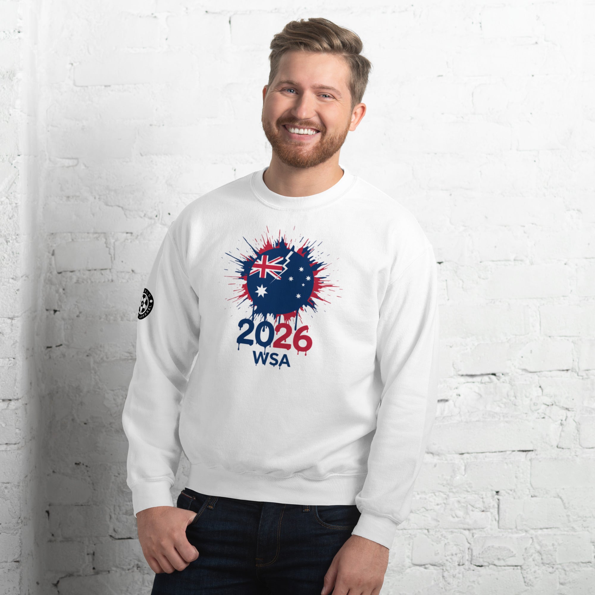 Man wearing a white sweatshirt with a design featuring the number 2026 and text, standing against a white brick wall.