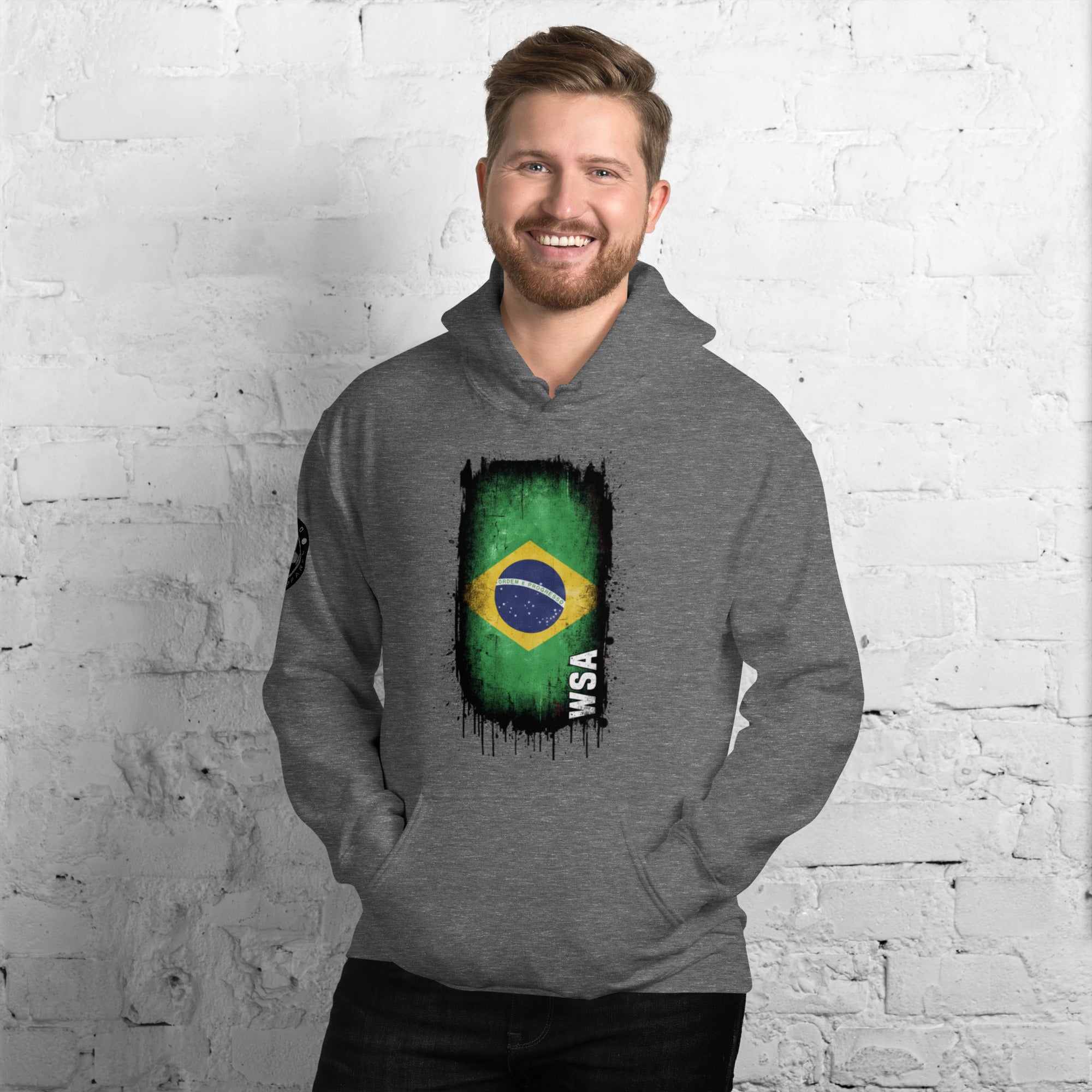 Man wearing a gray hoodie with a Brazilian flag design against a white brick wall.