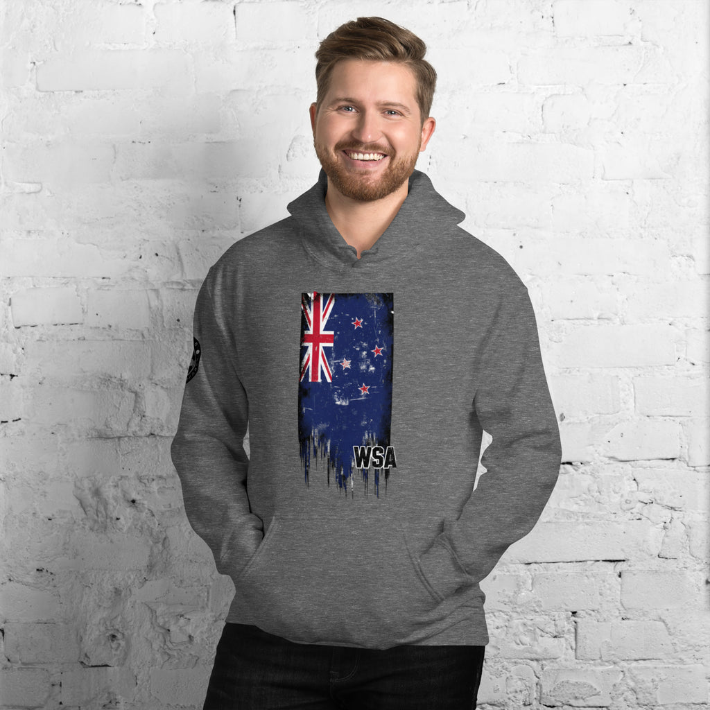 Man wearing a gray hoodie with a New Zealand flag design against a white brick wall.