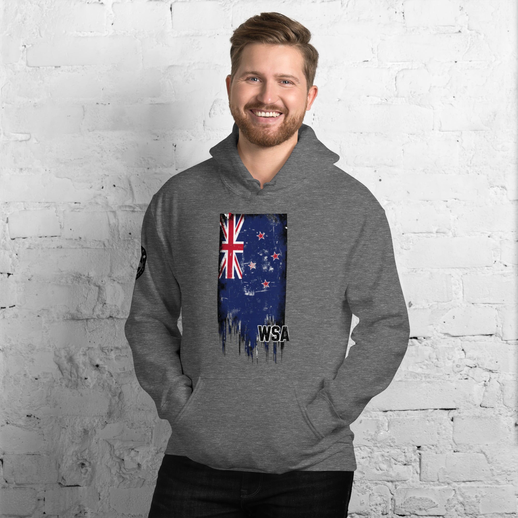 Man wearing a gray hoodie with a New Zealand flag design against a white brick wall.