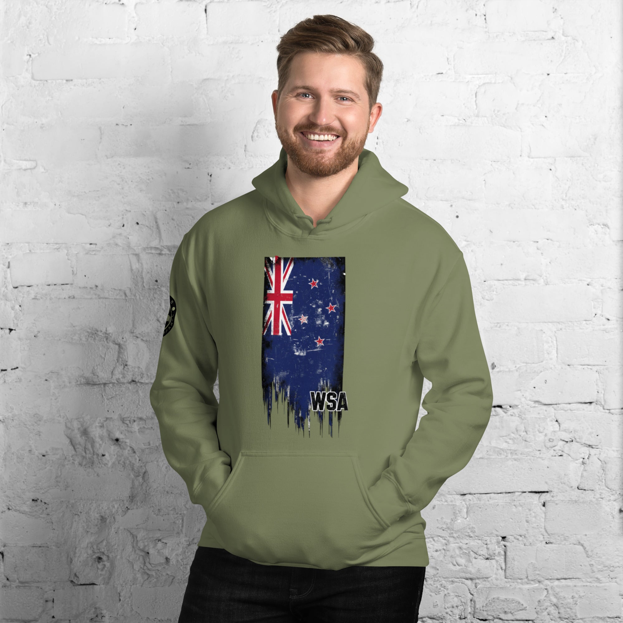 Man wearing a green hoodie with a New Zealand flag design against a white brick wall.