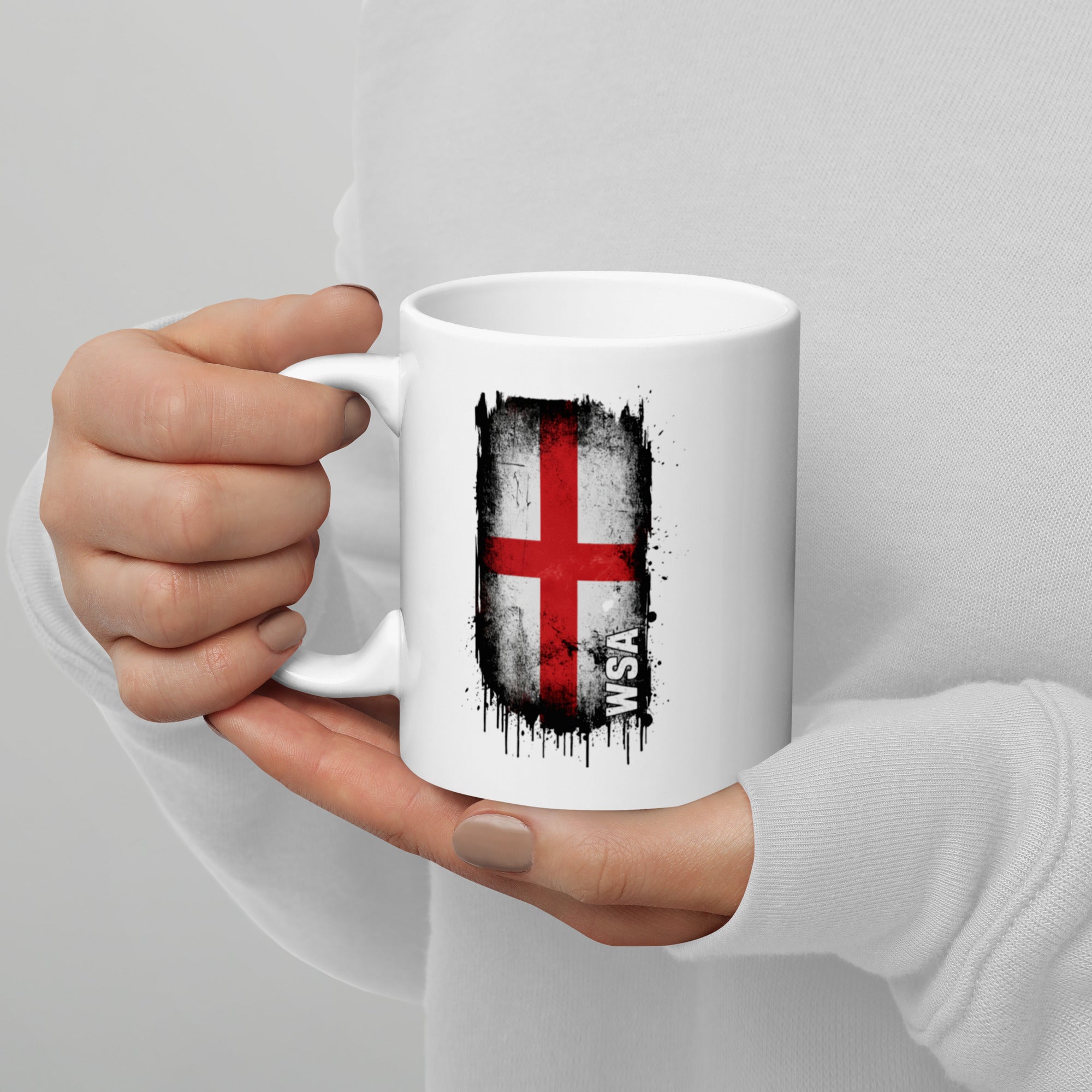 White mug with a red cross design held by a person against a gray background