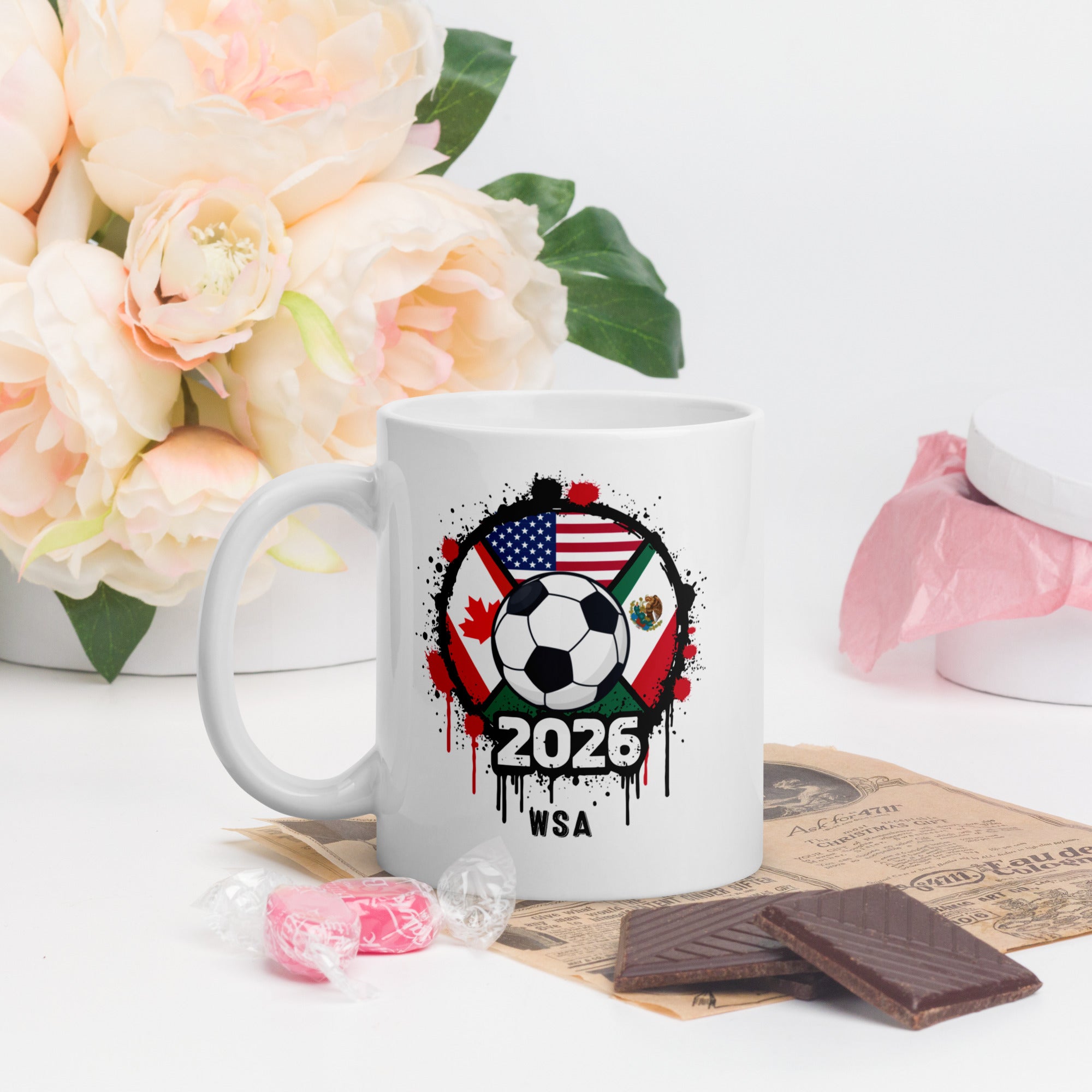 White mug with soccer-themed design featuring flags and '2026 WSA' text, placed on a surface with flowers and snacks.