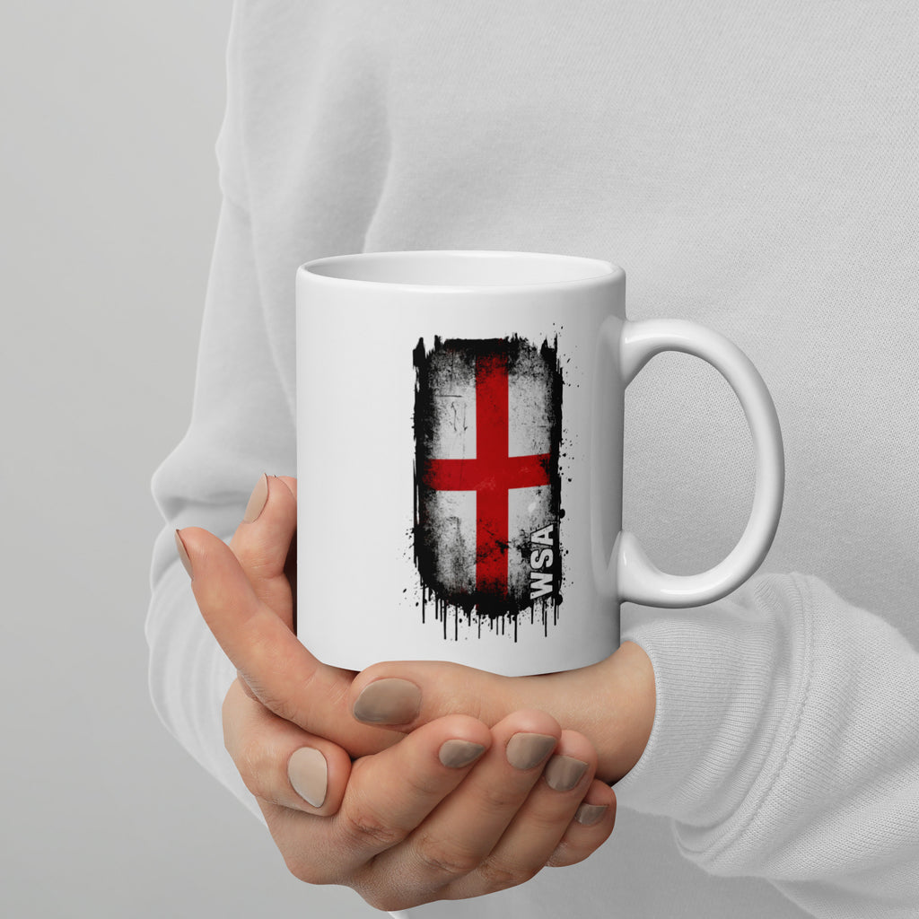White mug with a red cross design held by a person against a gray background