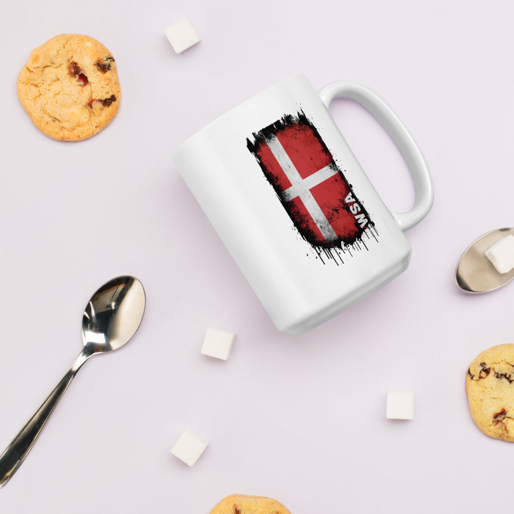 White mug with Danish flag design on a light surface with cookies and a spoon.