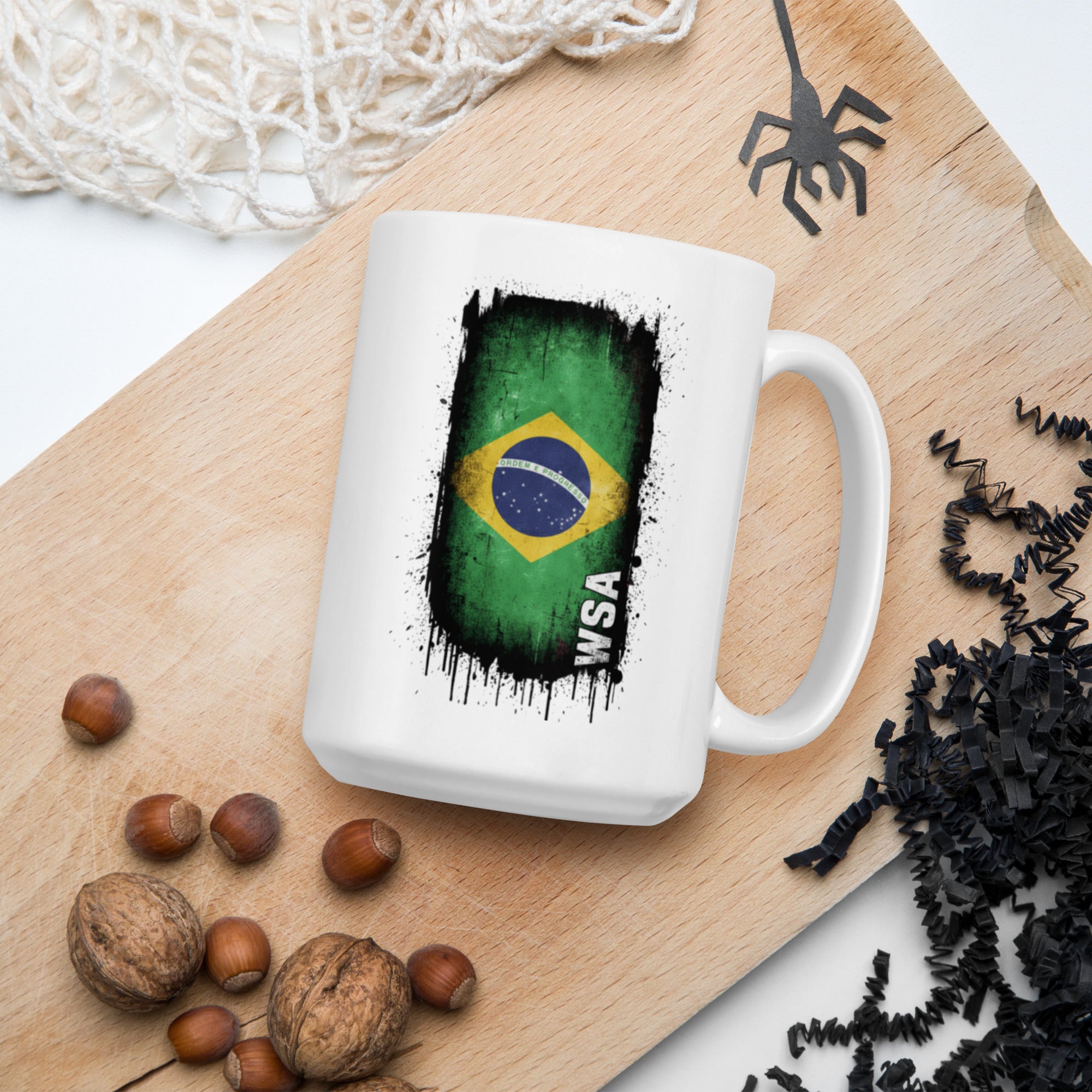 White mug with Brazilian flag design on a wooden board with nuts and spider decorations.