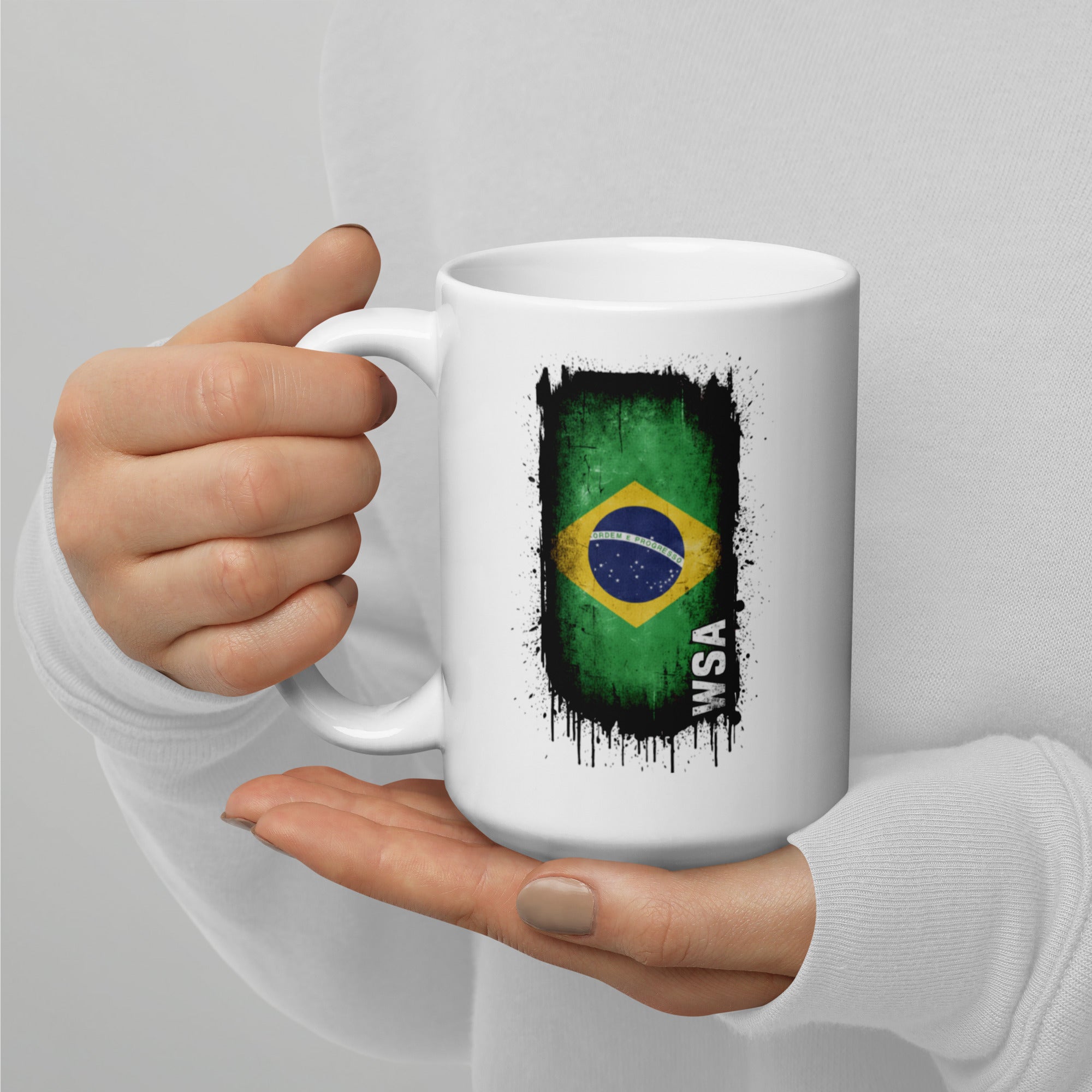 White mug with Brazilian flag design held by a hand on a light gray background