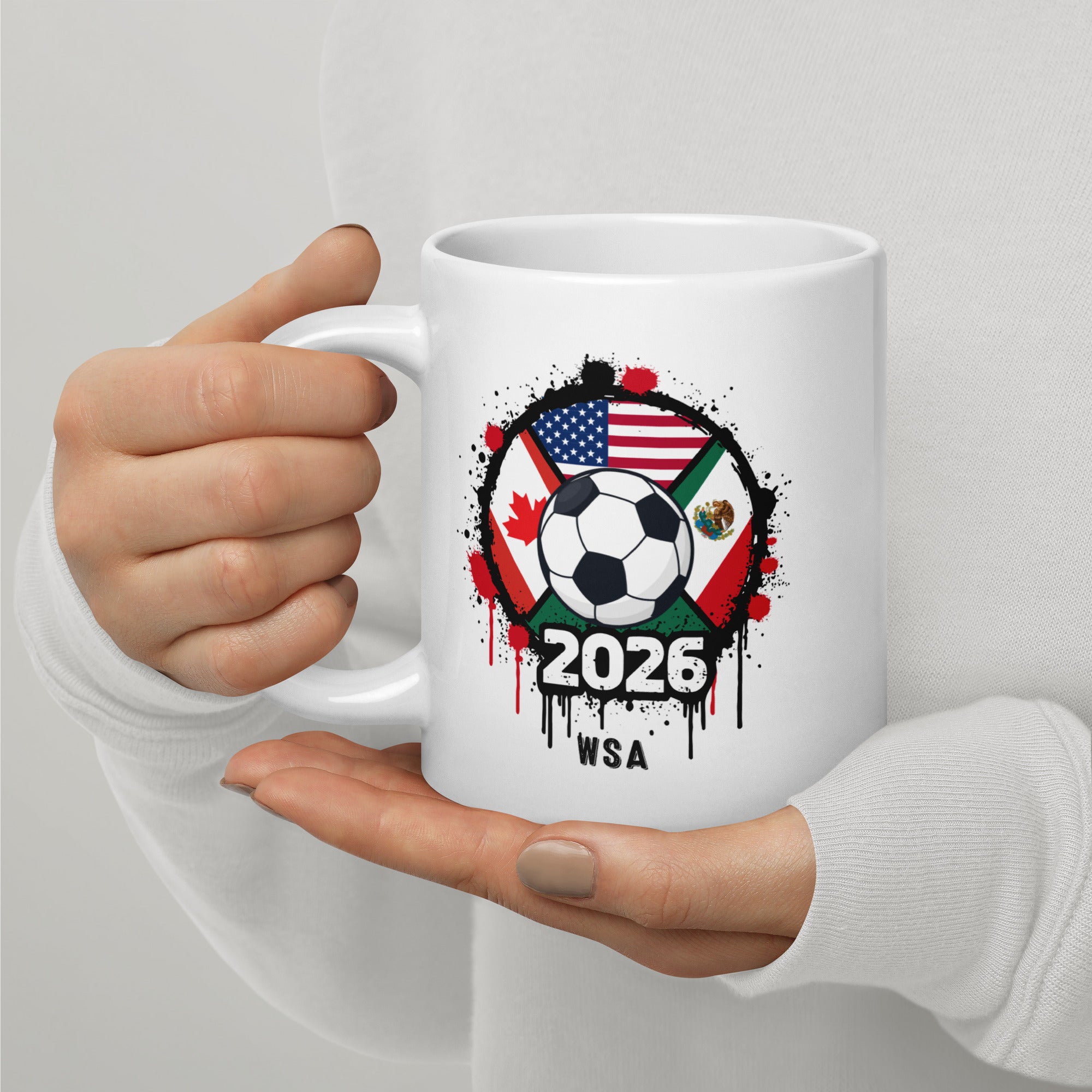 White mug with soccer ball design featuring flags and '2026 WSA' text, held by a person.