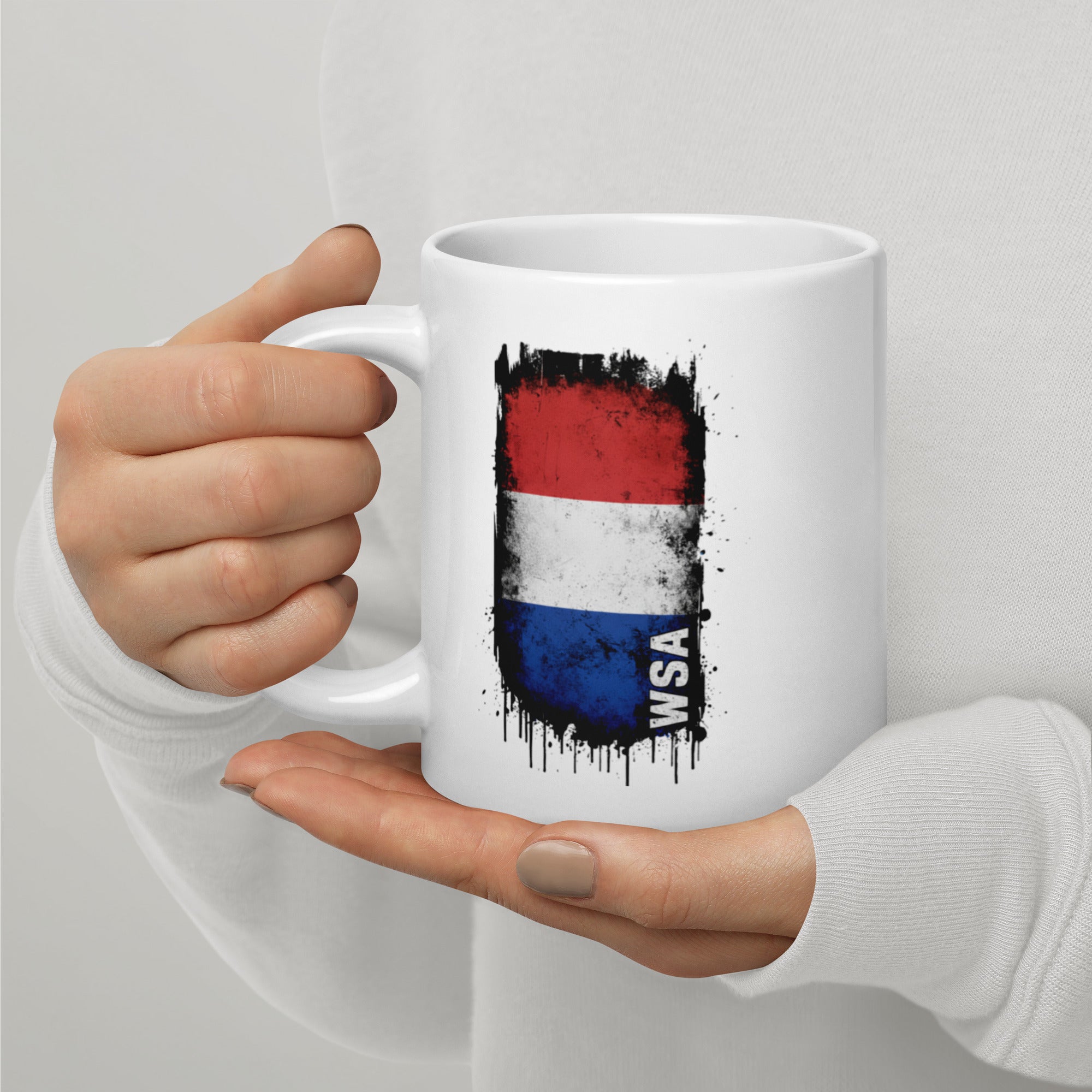 White mug with a flag design held by a person against a light gray background