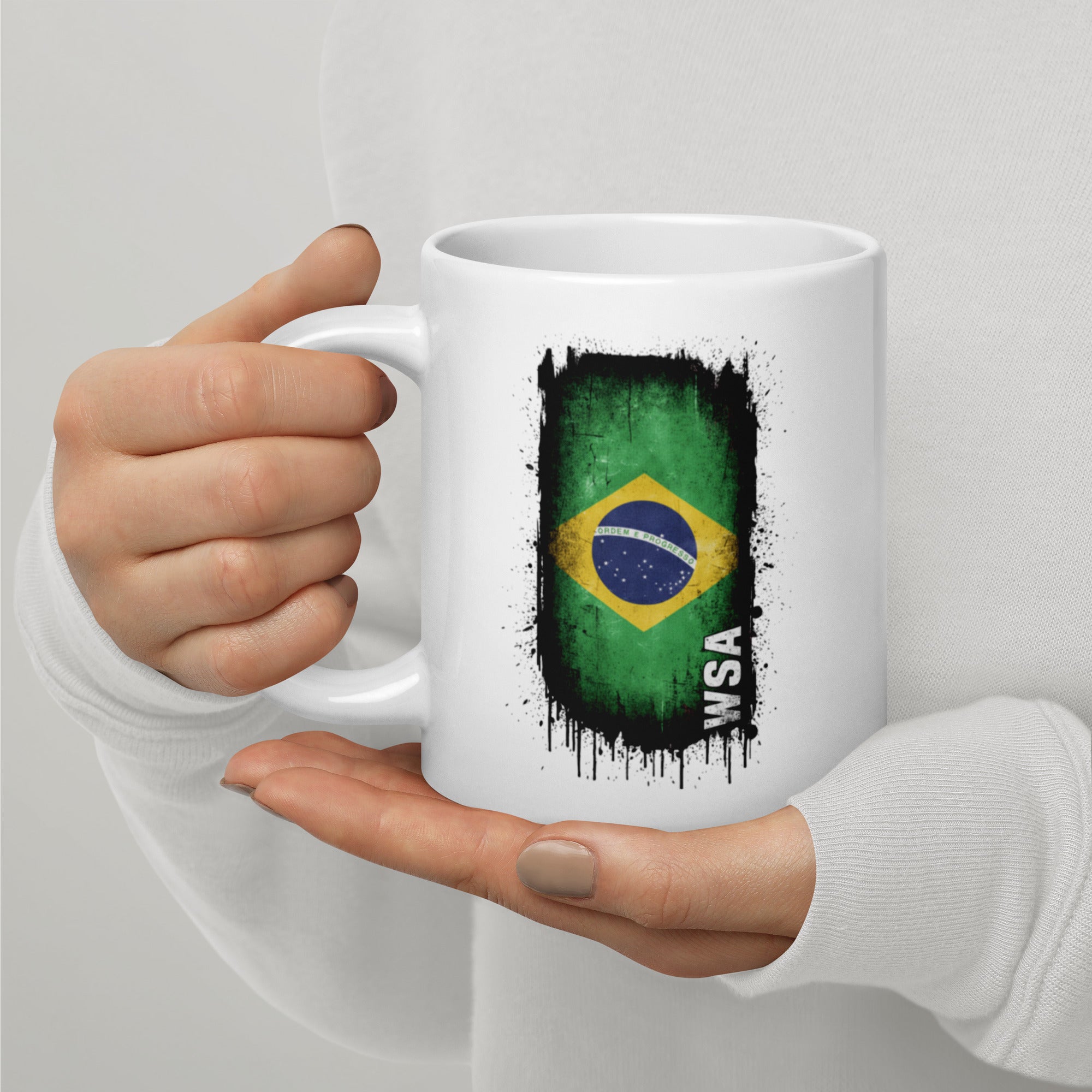 White mug with Brazilian flag design held by a person against a light gray background