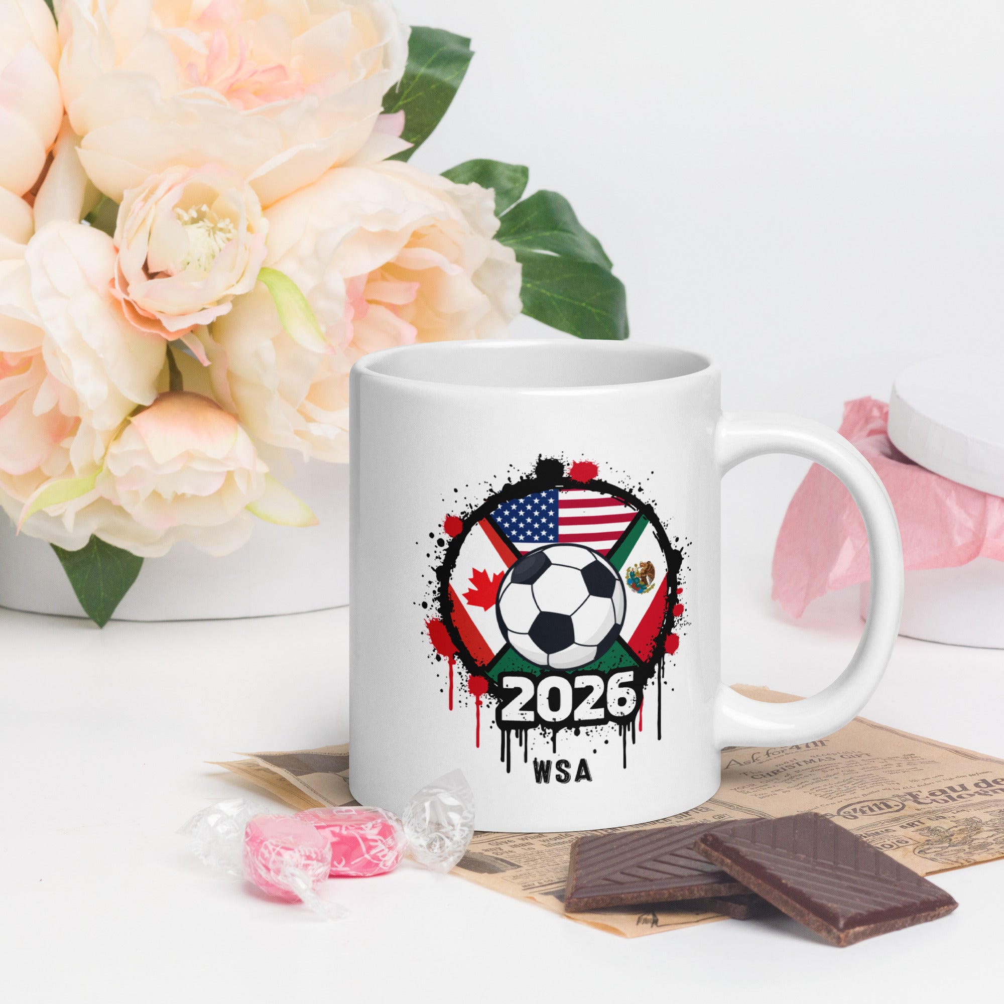 White mug with soccer ball design featuring flags and '2026 WSA' text, placed on a surface with flowers and snacks.