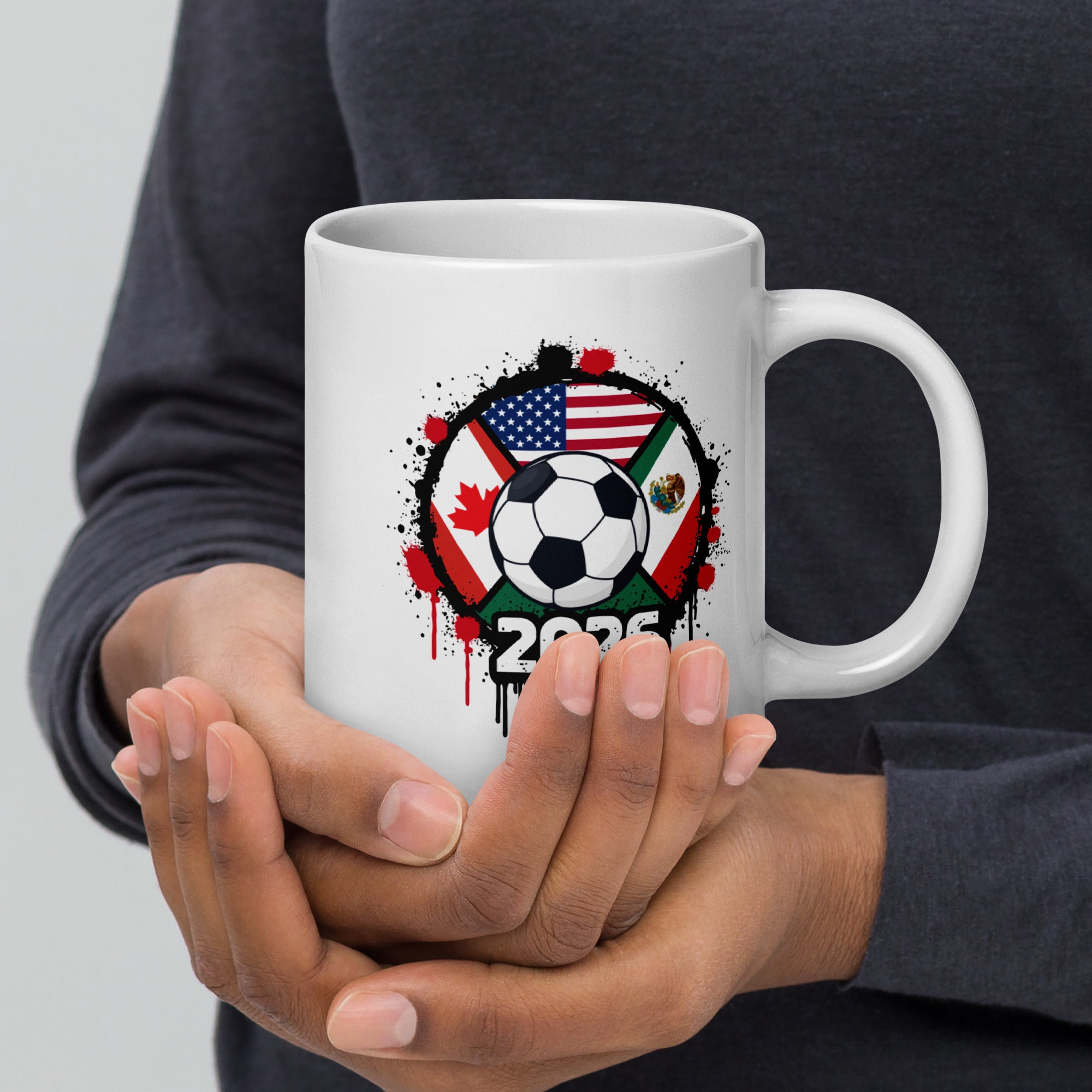 Person holding a white mug with a soccer ball and flags design, featuring the year 2026.