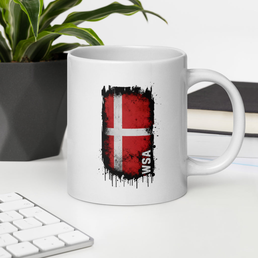 White mug with a red and black flag design on a desk with a keyboard and plant.