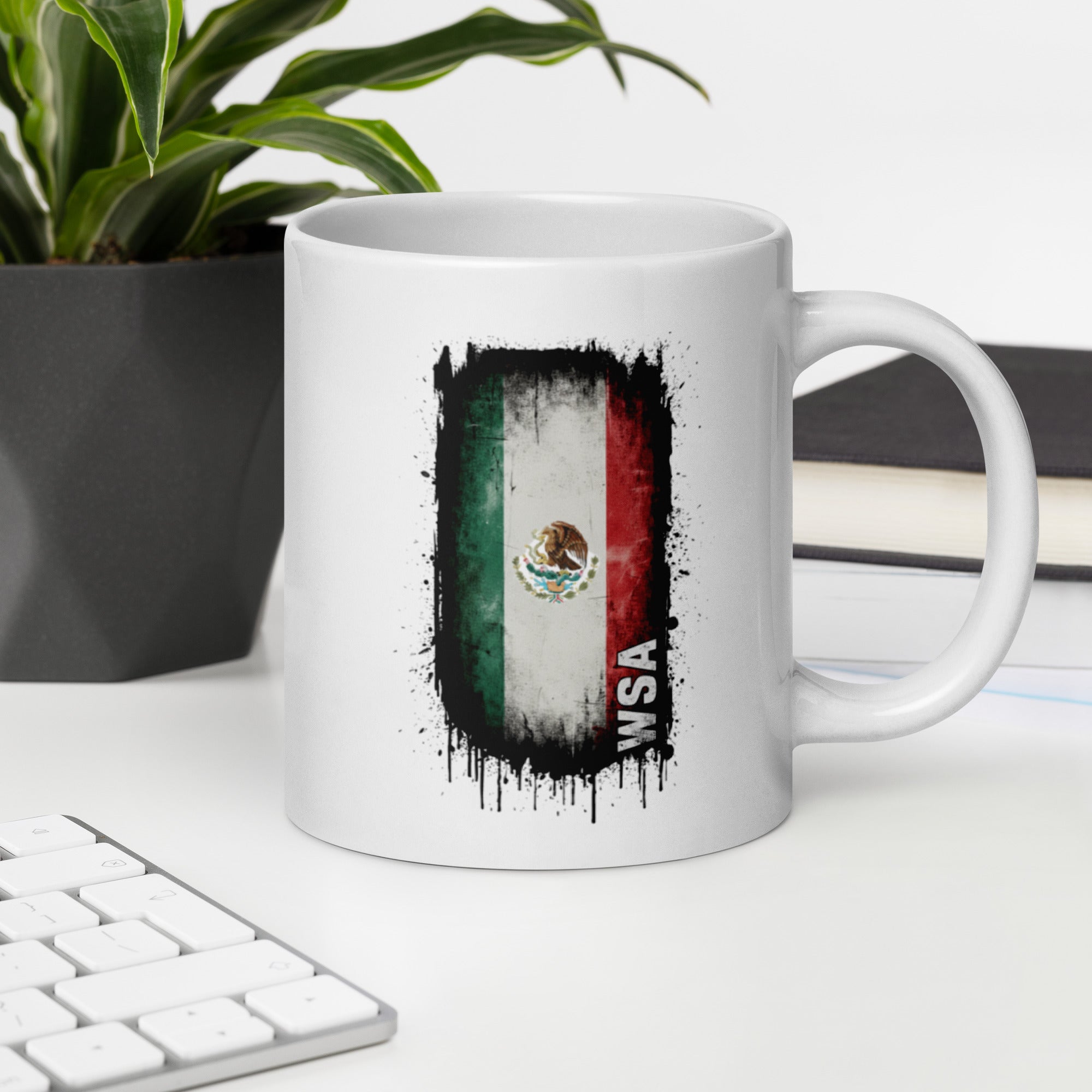 White mug with Mexican flag design on a desk with a keyboard and plant.