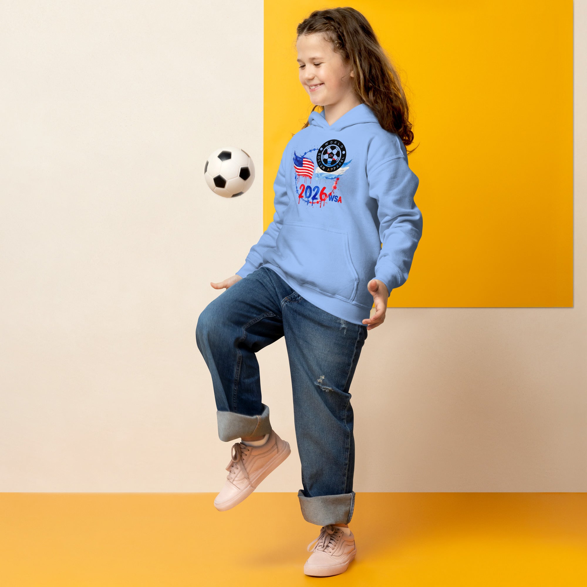 Child in a blue hoodie with sports-themed design, kicking a soccer ball against a yellow and beige background