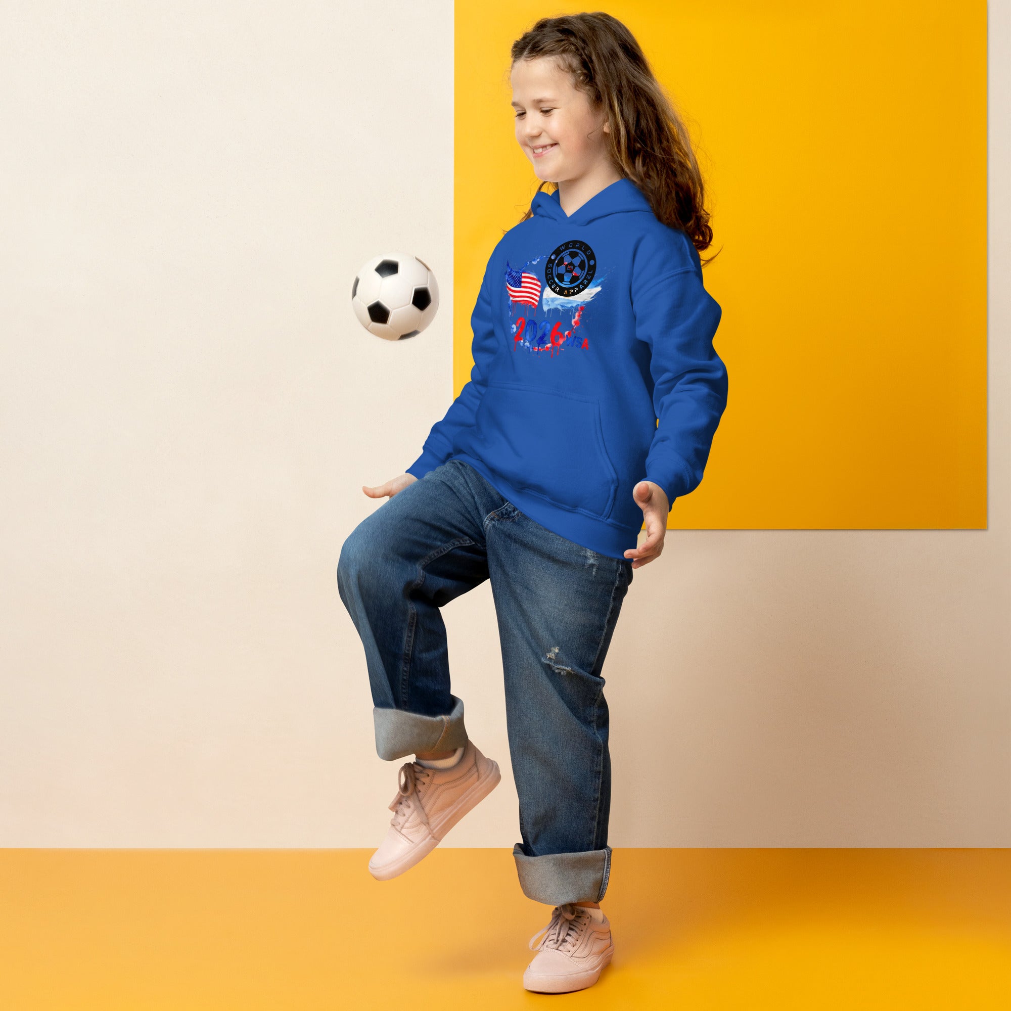 Child in a blue hoodie with a space-themed design, kicking a soccer ball against a yellow and beige background